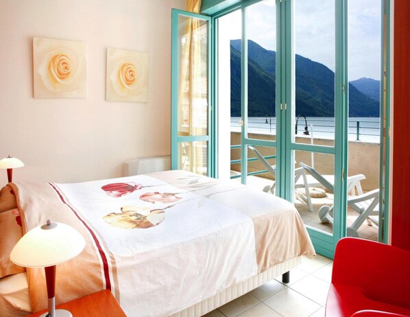 A bedroom with a bed, a nightstand, a lamp, a chair, and a view of a balcony with a mountain and water.