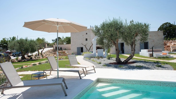 Ein Poolbereich mit Liegestühlen und Sonnenschirm, umgeben von einem angelegten Garten und einem Gebäude mit mehreren Eingängen.