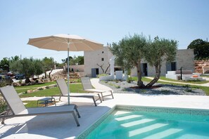 A poolside area with lounge chairs and an umbrella, surrounded by a landscaped garden and a building with multiple doors. - Tower House in Puglia With Pool and Charm (Cisternino)