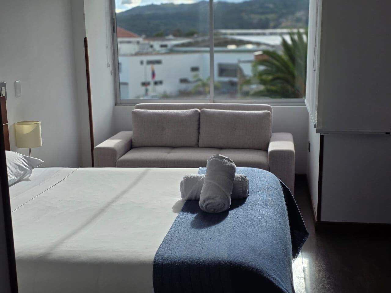 A hotel room with a bed, a sofa, a window with a view of buildings and hills, and a lamp.