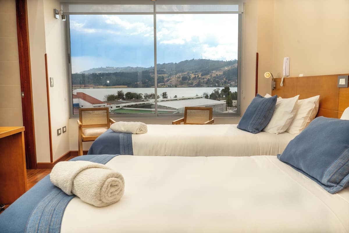 A hotel room with two beds, a large window with a view of a lake and hills, and a wooden headboard.