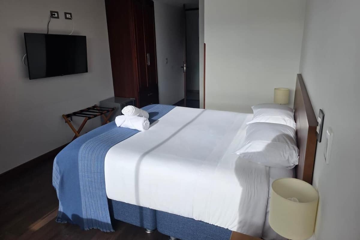 A hotel room with a bed, a TV mounted on the wall, a wardrobe, and a small table with towels.