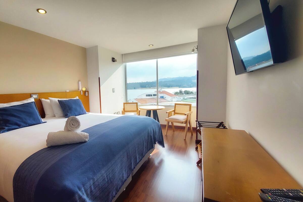 A hotel room with a large bed, a desk, a chair, a TV, and a view of a body of water.