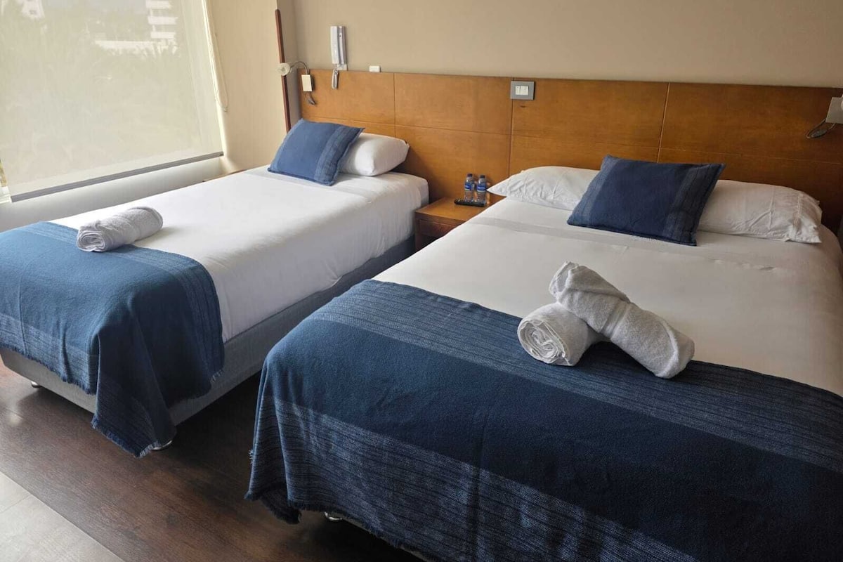 Two beds with blue and white bedding, a wooden headboard, a nightstand with a lamp, and a window with blinds.