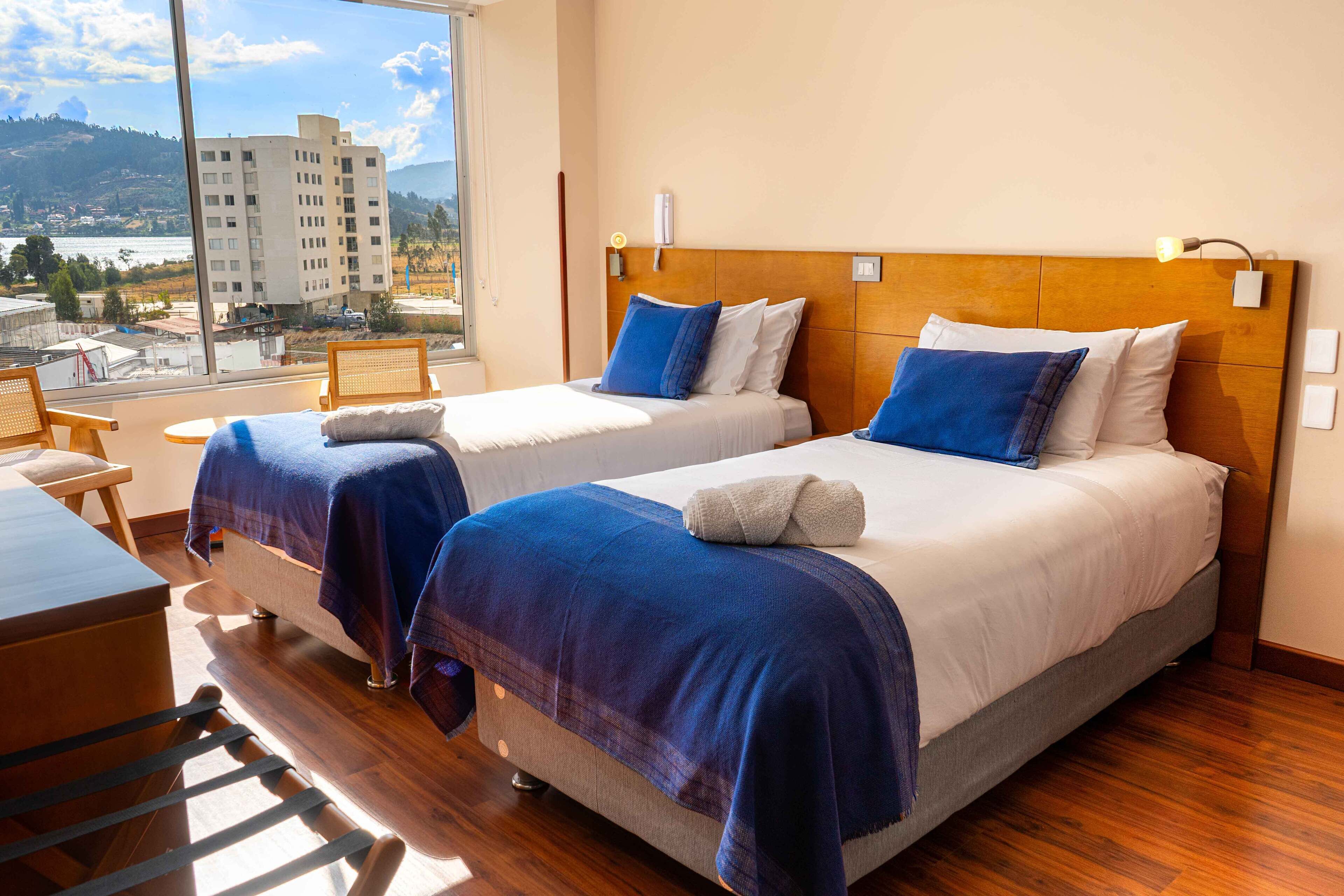 Habitación de hotel con dos camas, suelo de madera, vistas a un edificio y colinas, y una pequeña mesa con sillas.