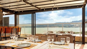 Un restaurant où les tables sont dressées pour le service, de grandes fenêtres offrant une vue sur un lac et des collines, et un décor intérieur moderne.