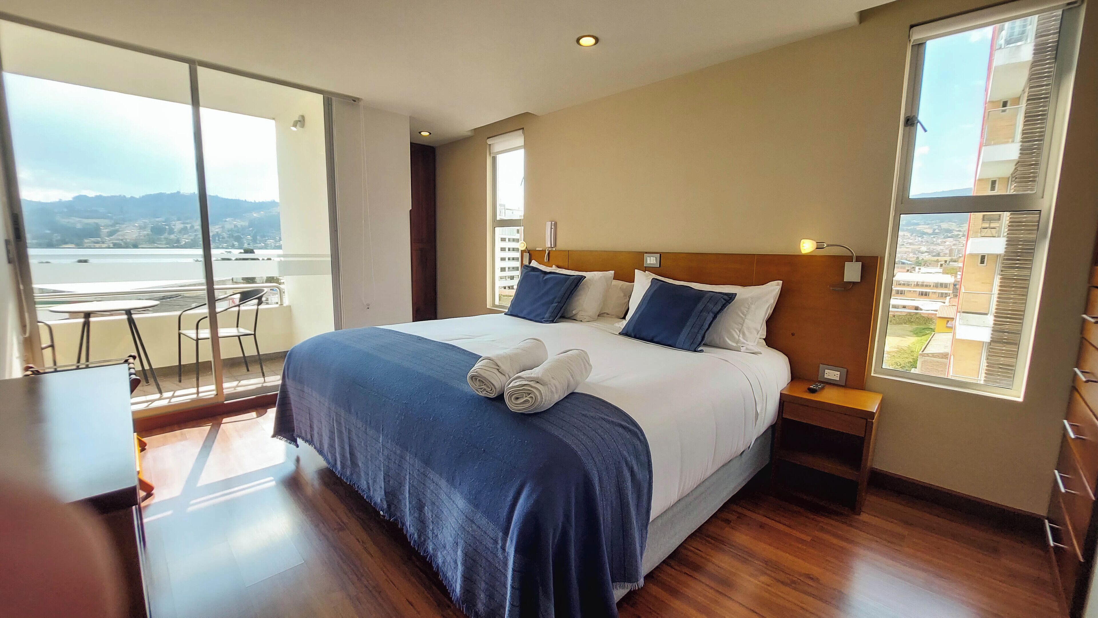 Habitación de hotel con una cama grande, una mesita de noche, un escritorio con una silla y un balcón con mesa y sillas.