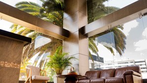Un salon moderne avec un canapé en cuir, une chaise en bois et une table basse. De grandes fenêtres laissent entrer la lumière naturelle et offrent une vue sur les palmiers.