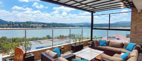 A covered outdoor seating area with wicker furniture and a view of a lake and distant hills.
