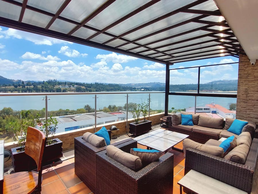 Un espacio de estar techado al aire libre con mobiliario de mimbre y vistas a un lago y colinas distantes.