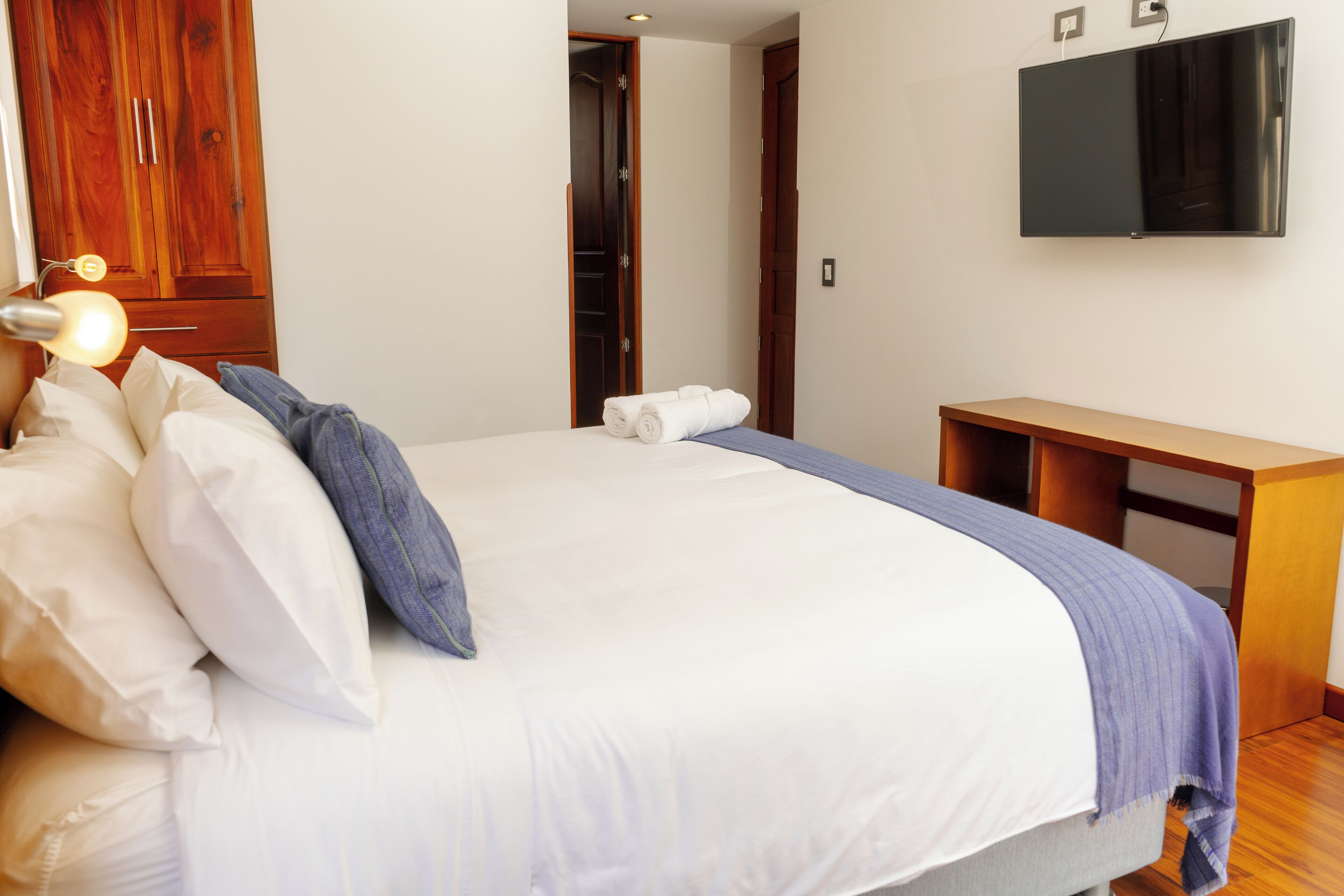 Una cama bien hecha con ropa de cama blanca y azul, una mesita de noche de madera, un televisor de pantalla plana montado en la pared y un armario de madera.
