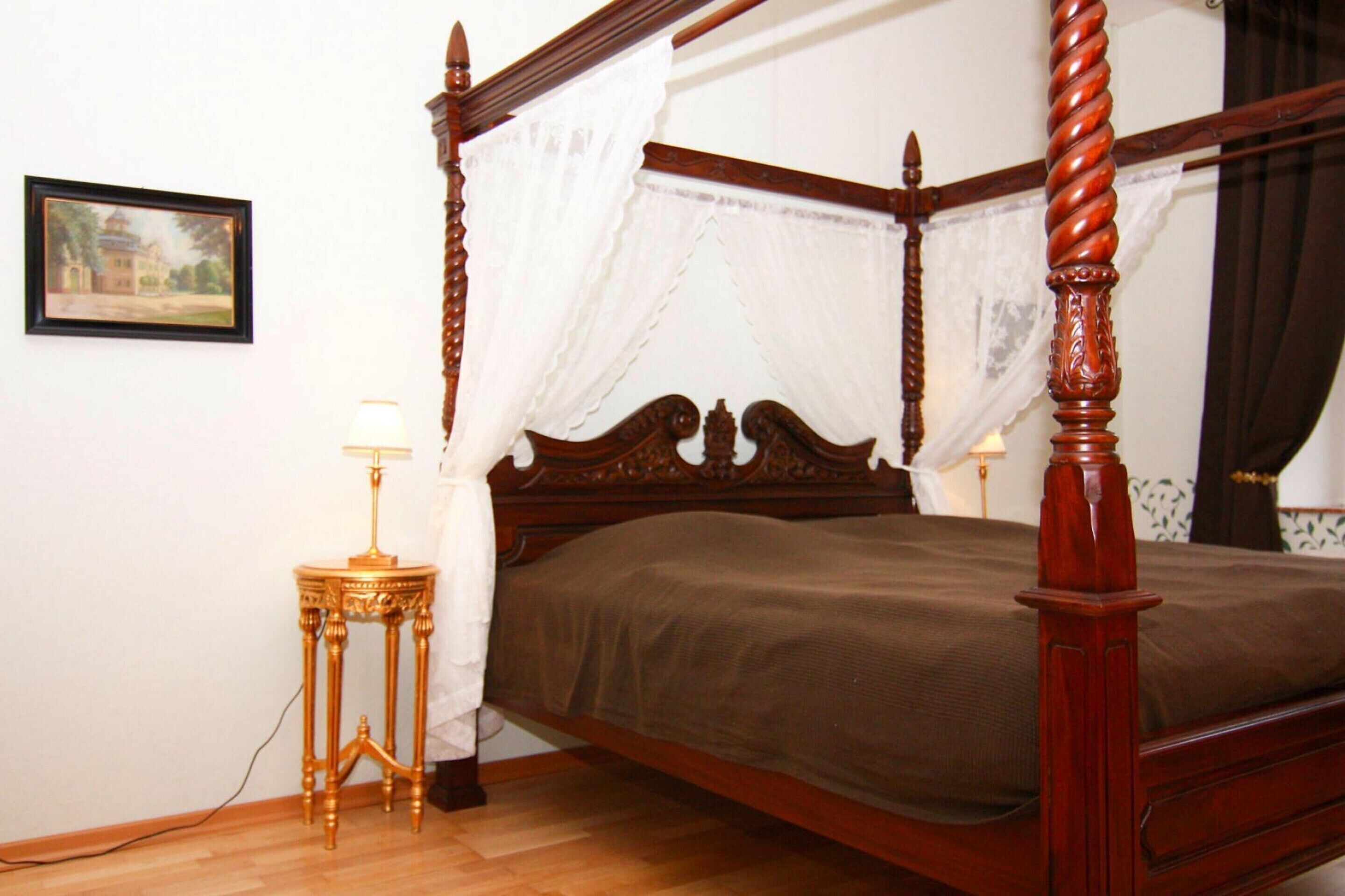 A four-poster bed with a wooden frame and white curtains, a bedside table with a lamp, and a framed picture on the wall.