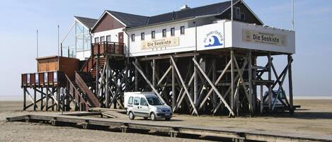 A wooden pier structure with a restaurant named "Die Seekiste" on stilts over a sandy beach.