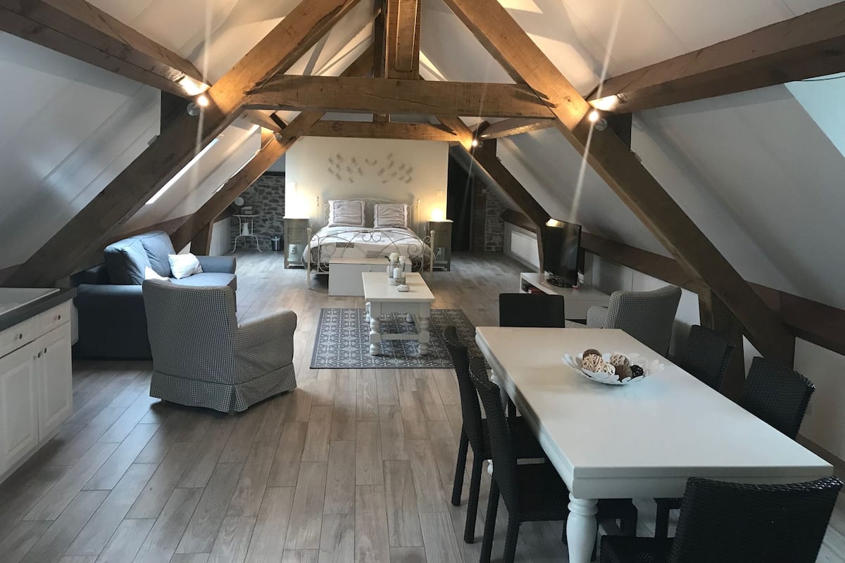 A spacious attic bedroom with a large bed, a sitting area with a sofa and chairs, and a dining table with a bowl of fruit.
