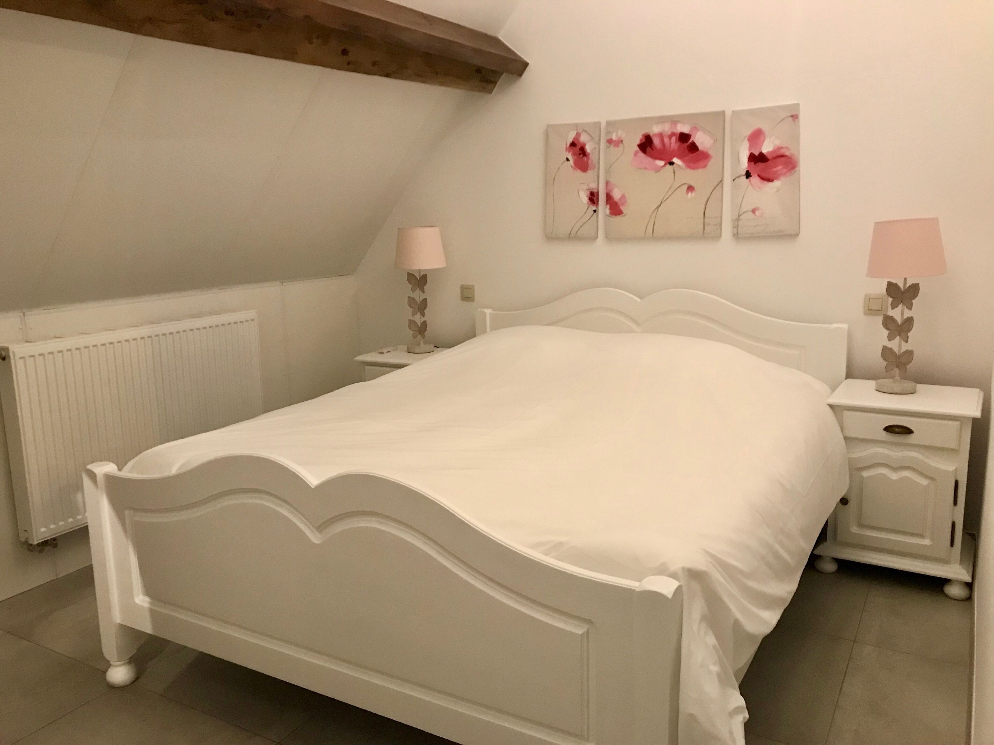 A white bedroom with a bed, two bedside tables, a radiator, and three wall-mounted flower paintings.