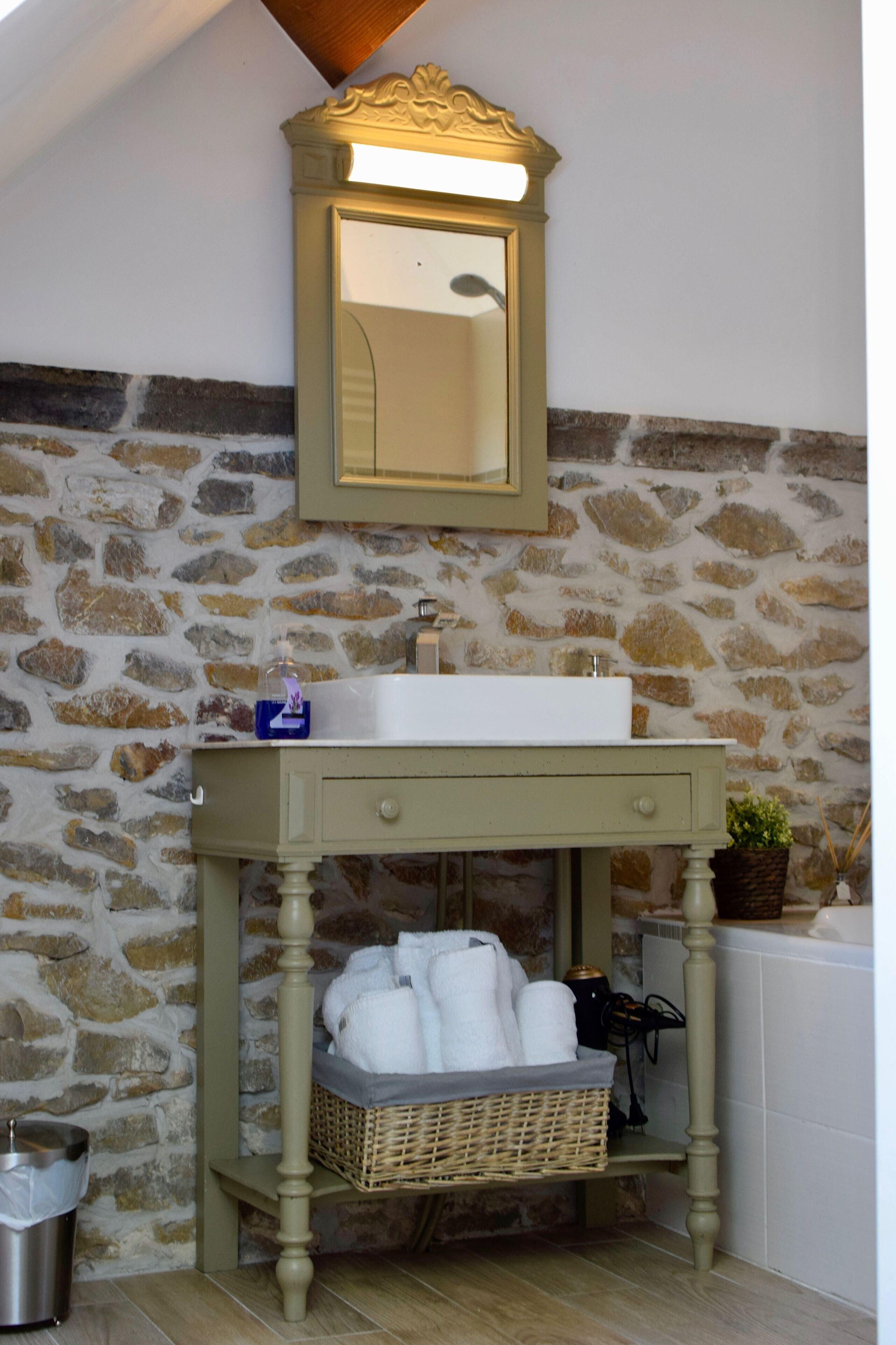 A bathroom with a stone wall, a sink, a mirror, a basket with towels, and a trash can.