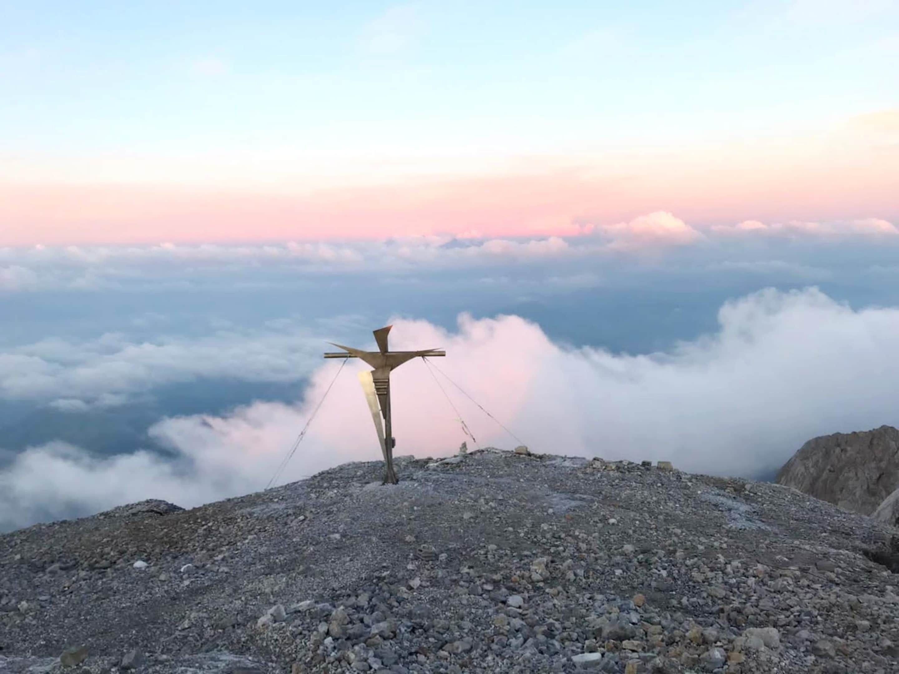 A cross on a mountain peak with a view of clouds below.