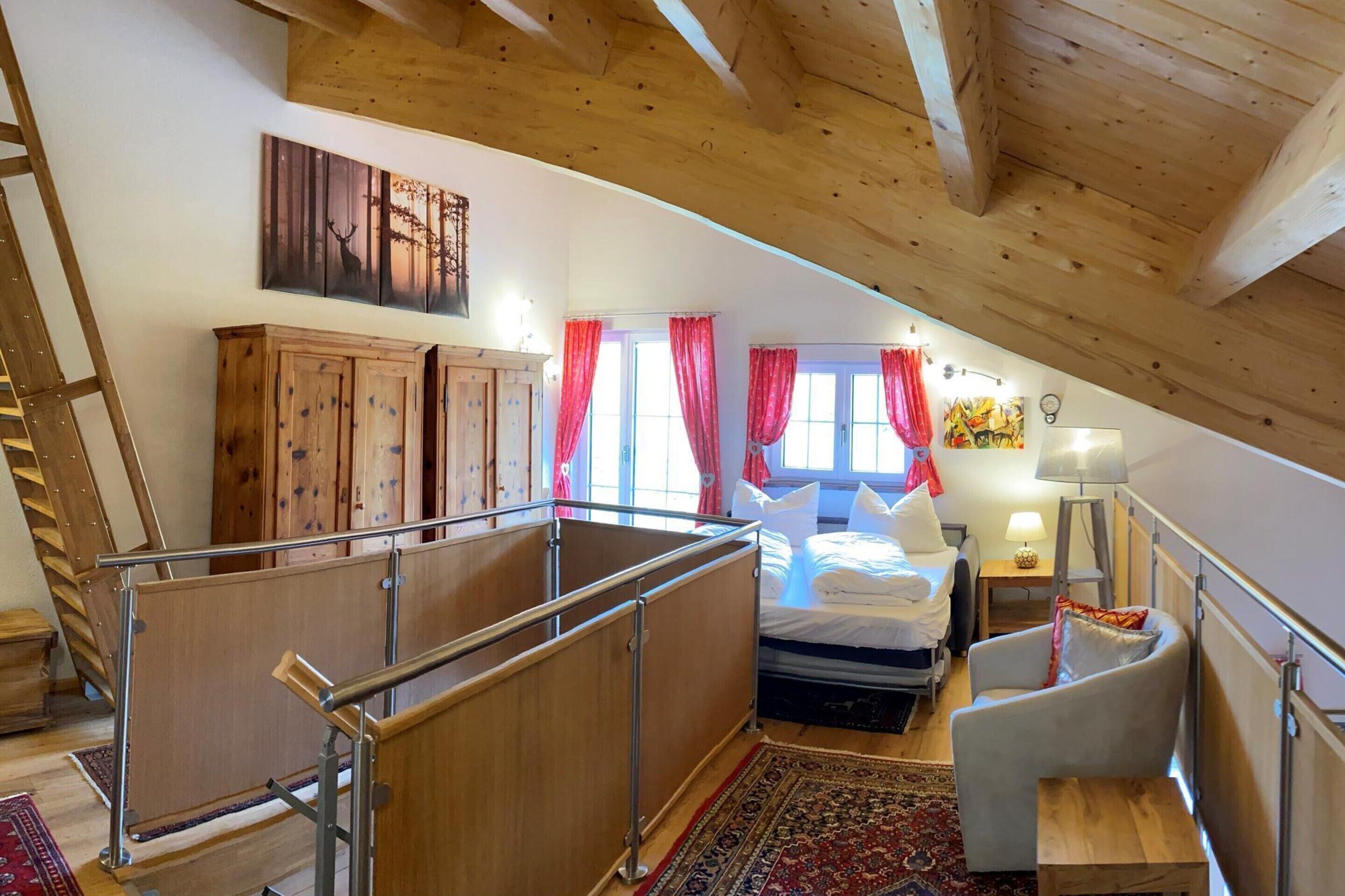 Une chambre mansardée chaleureuse avec un plafond en bois, un lit recouvert de draps blancs, une chaise, une table de chevet et un rideau rouge.