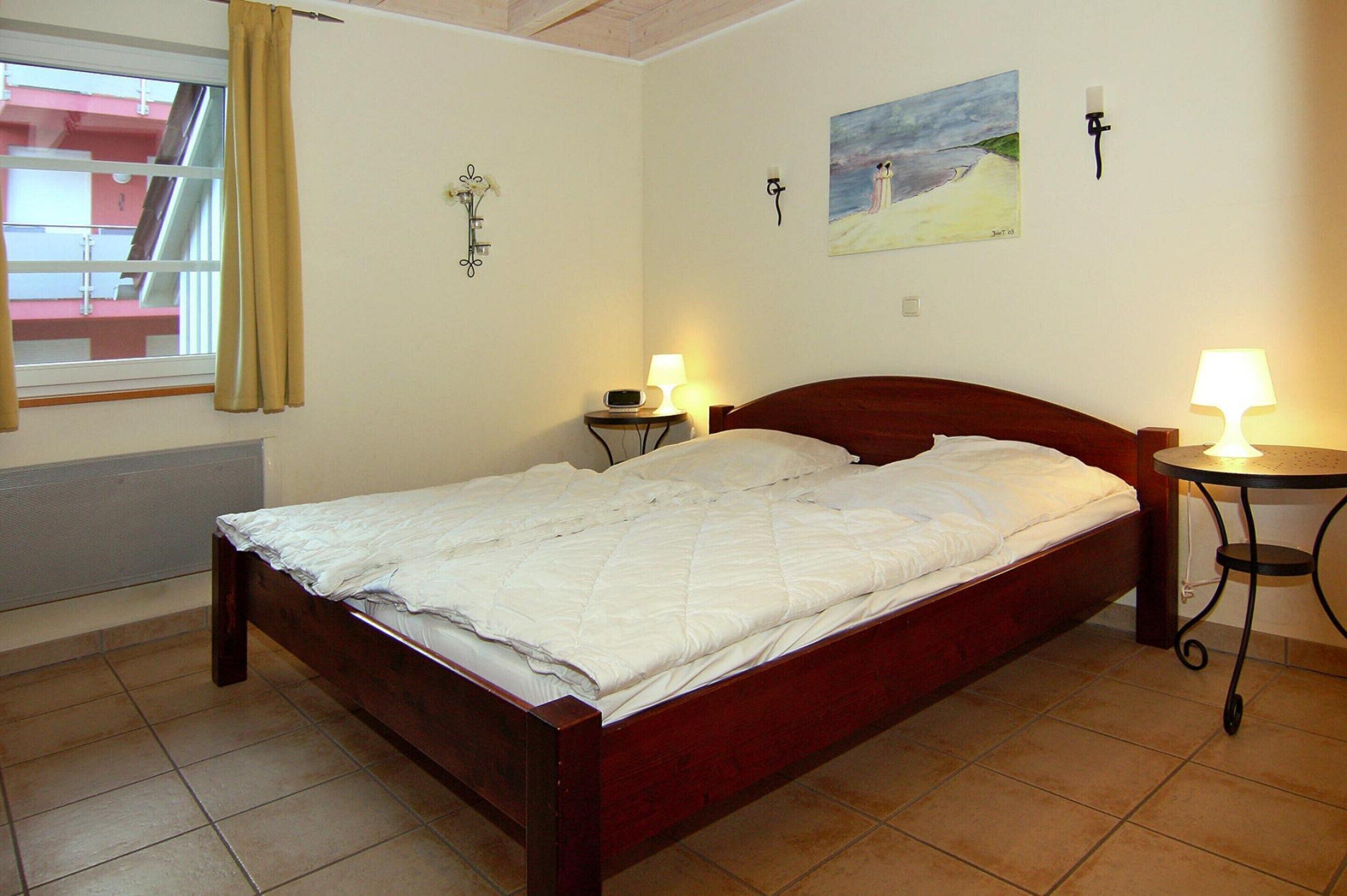 A bedroom with a wooden bed, two bedside tables with lamps, a window with curtains, and a painting on the wall.