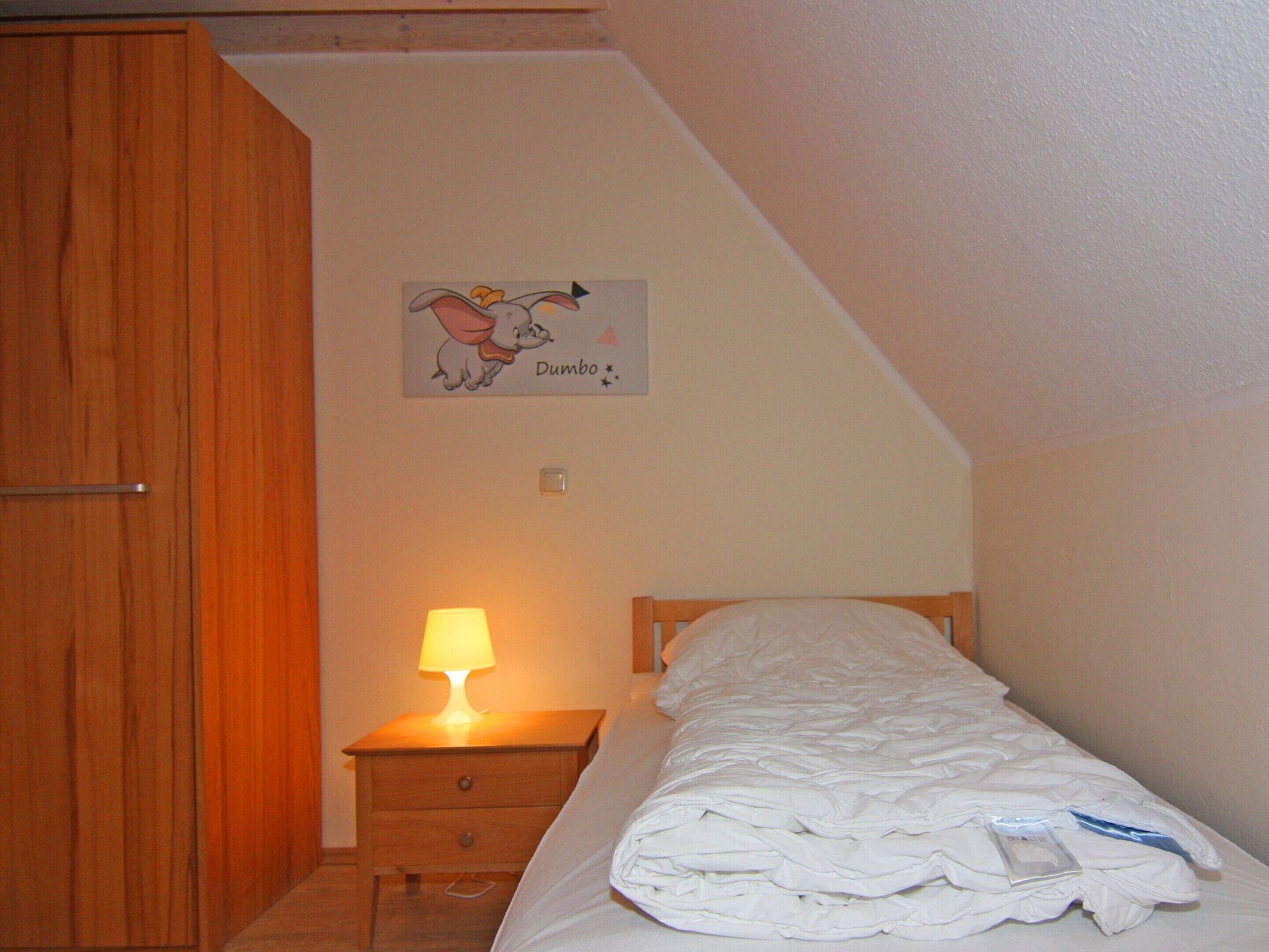 Une chambre avec une armoire en bois, une table de chevet avec une lampe, un lit recouvert d’une couette blanche et une décoration murale représentant un personnage de dessin animé.