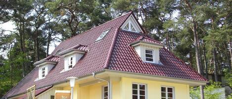 Une maison à deux étages, avec un toit recouvert de tuiles rouges, une véranda avec des chaises et un petit jardin.