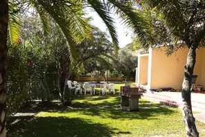 An outdoor seating area with white plastic chairs and tables, surrounded by lush greenery and palm trees. - Apartment on Costa Rei Near White Beaches (Muravera)