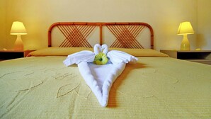 Una cama bien hecha con una toalla doblada en forma de cisne y una pequeña flor sobre la almohada. Hay dos lámparas de noche a ambos lados de la cama.
