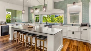 A modern kitchen with a large island, bar stools, and a view of greenery outside. - koolkondo 2606 Halalu Main house (Koloa)