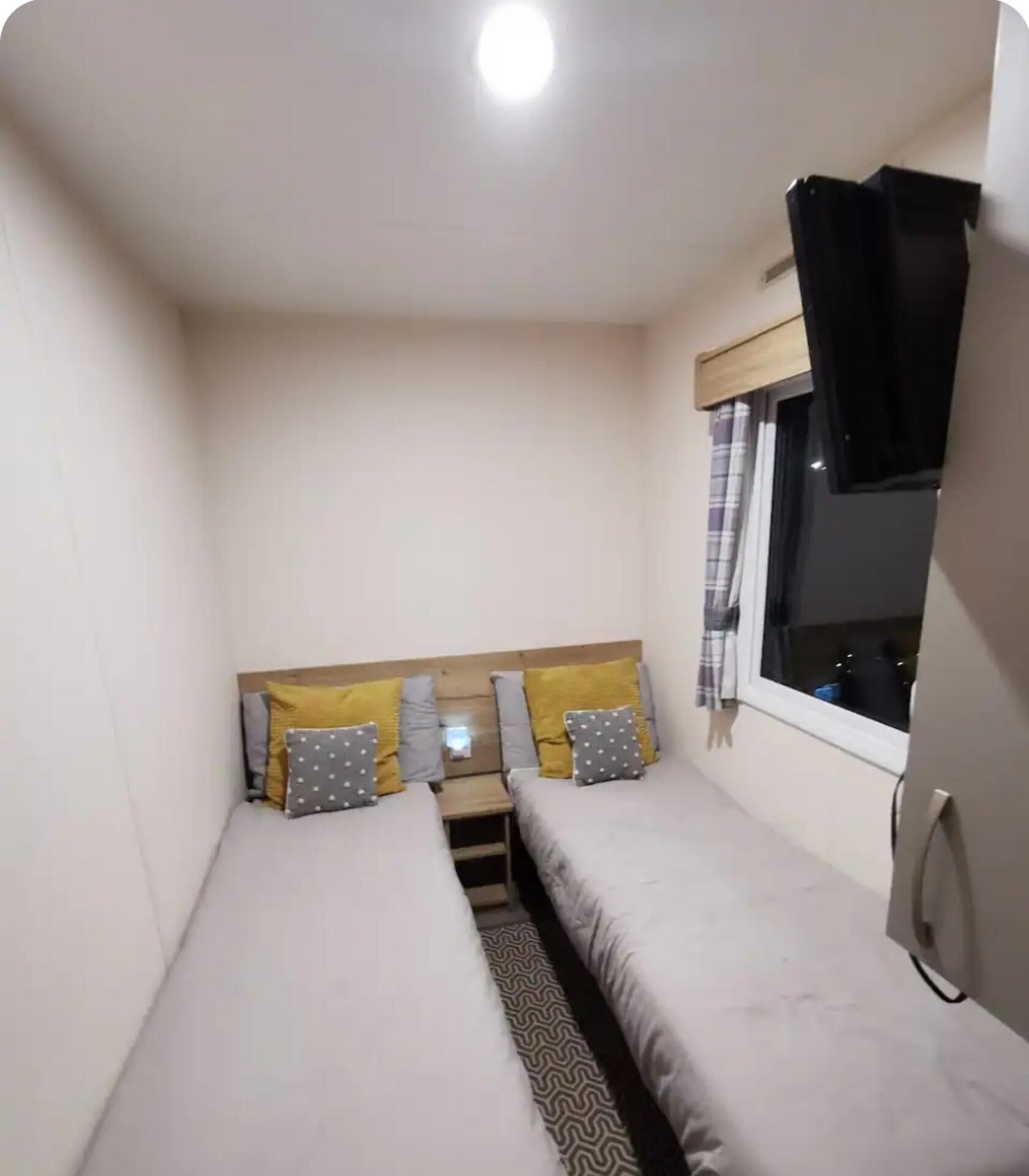 A small, single-room accommodation with two single beds, a window with curtains, and a ceiling light.