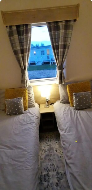 A small room with two beds, a window with curtains, a nightstand with a lamp, and a view of a house outside. - Cosy Caravan Retreat - Hot Tub and Free Parking (Lincoln)