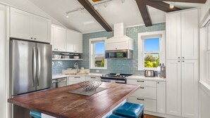A modern kitchen with white cabinets, a stainless steel refrigerator, a central island with stools, and a view of greenery through windows. - koolkondo 2606 Halalu Guest house (Koloa)