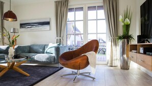 Ein modernes Wohnzimmer mit einer Couch, einem braunen Sessel, einem Glastisch, einem Fernseher und Blick auf ein Haus durch das Fenster.