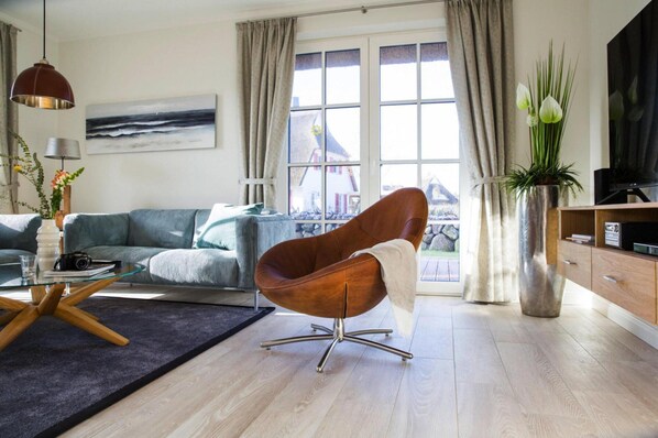 Un salon moderne avec un canapé, un fauteuil marron, une table basse en verre, un téléviseur et une vue sur une maison par la fenêtre.