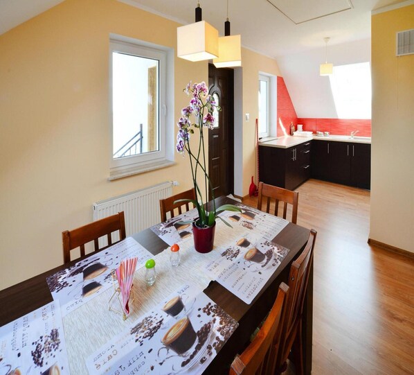 A dining area with a table set for two, featuring a menu with coffee images. There are chairs, a potted orchid, and a vase with flowers. The room has wooden flooring, a window, and a view of the kitchen in the background. - Holiday Apartment, 2 Bedrooms, Garden, Bicycles (Kołobrzeg)