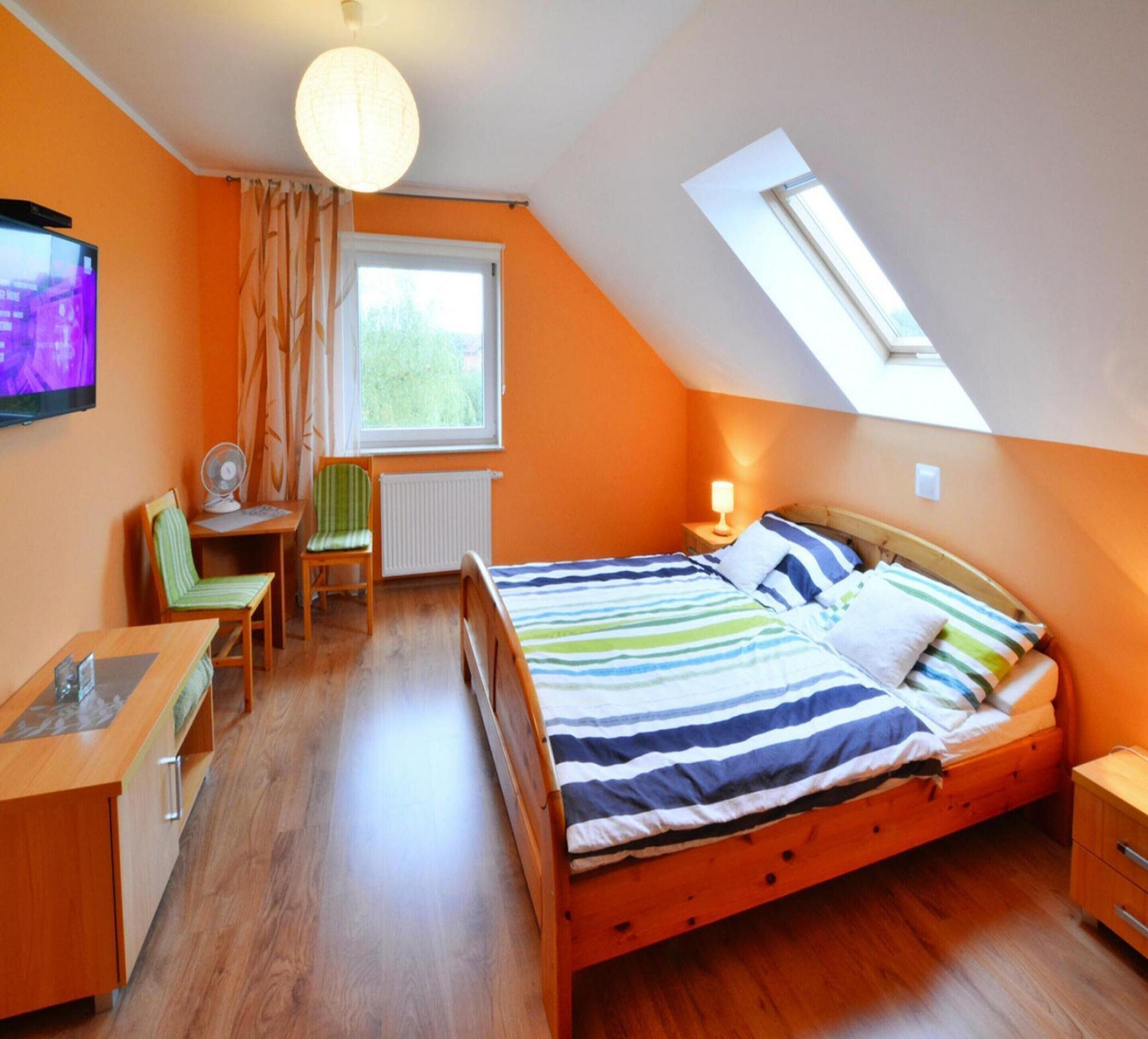 A bedroom with a sloped ceiling, a bed with striped bedding, a desk with a chair, a television, and a window with curtains.