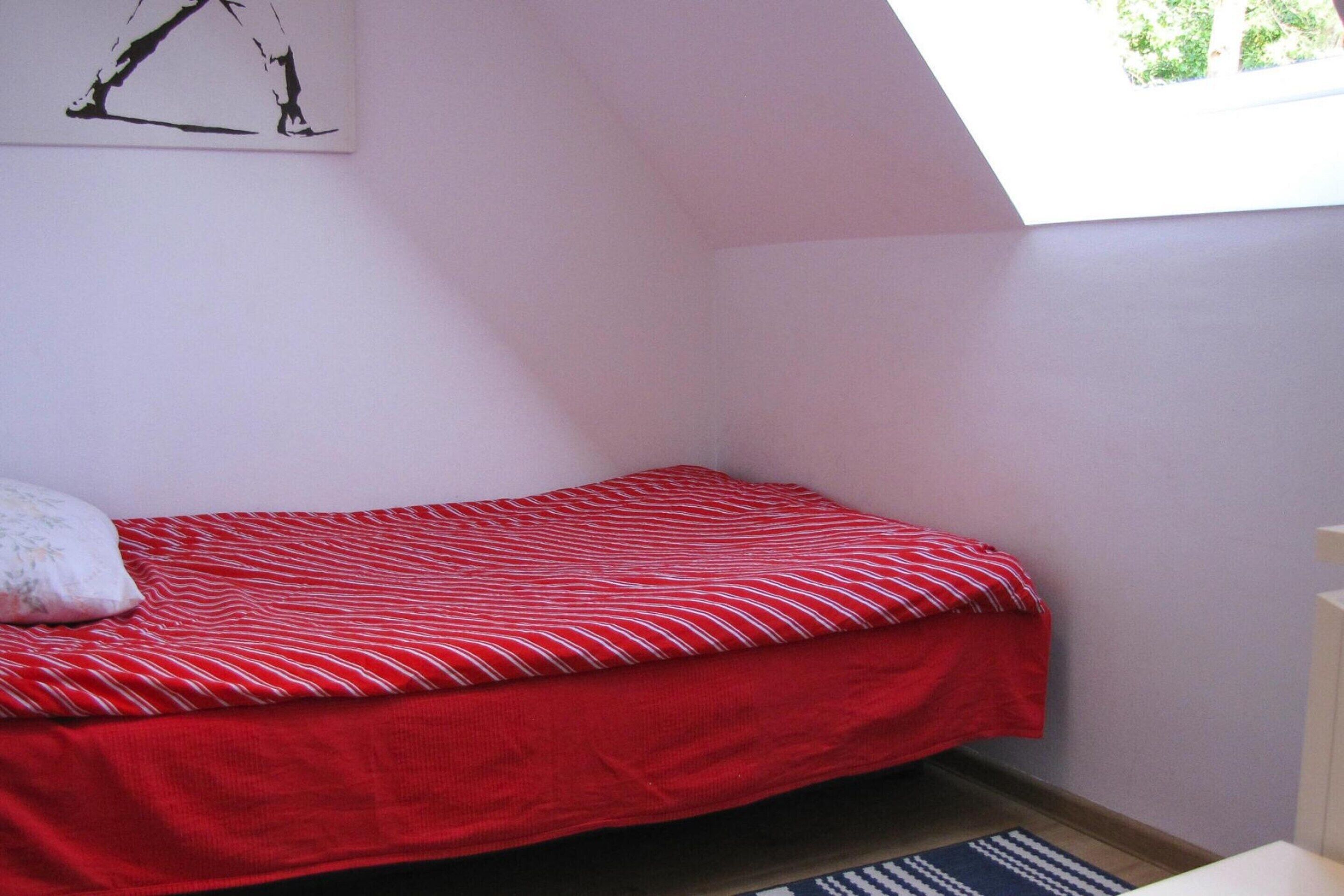 A small, single-bed room with a red-striped bedspread, a white pillow, and a window with a view of greenery.
