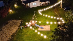 Une cour arrière avec une aire de jeux, des guirlandes lumineuses et une maison en arrière-plan.