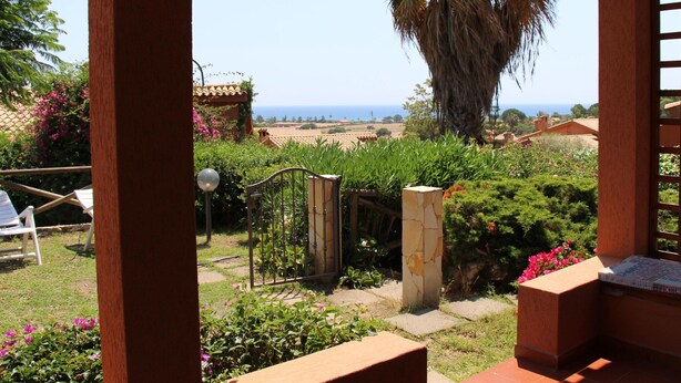 A patio with a view of the sea, a palm tree, and a garden with flowers and shrubs. - Apartment in Costa Rei Near White Sandy Beach (Muravera)