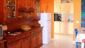 A kitchen with wooden cabinets, a white refrigerator, and a dining area with a blue chair. - Apartment in Costa Rei Near White Sandy Beach (Muravera)