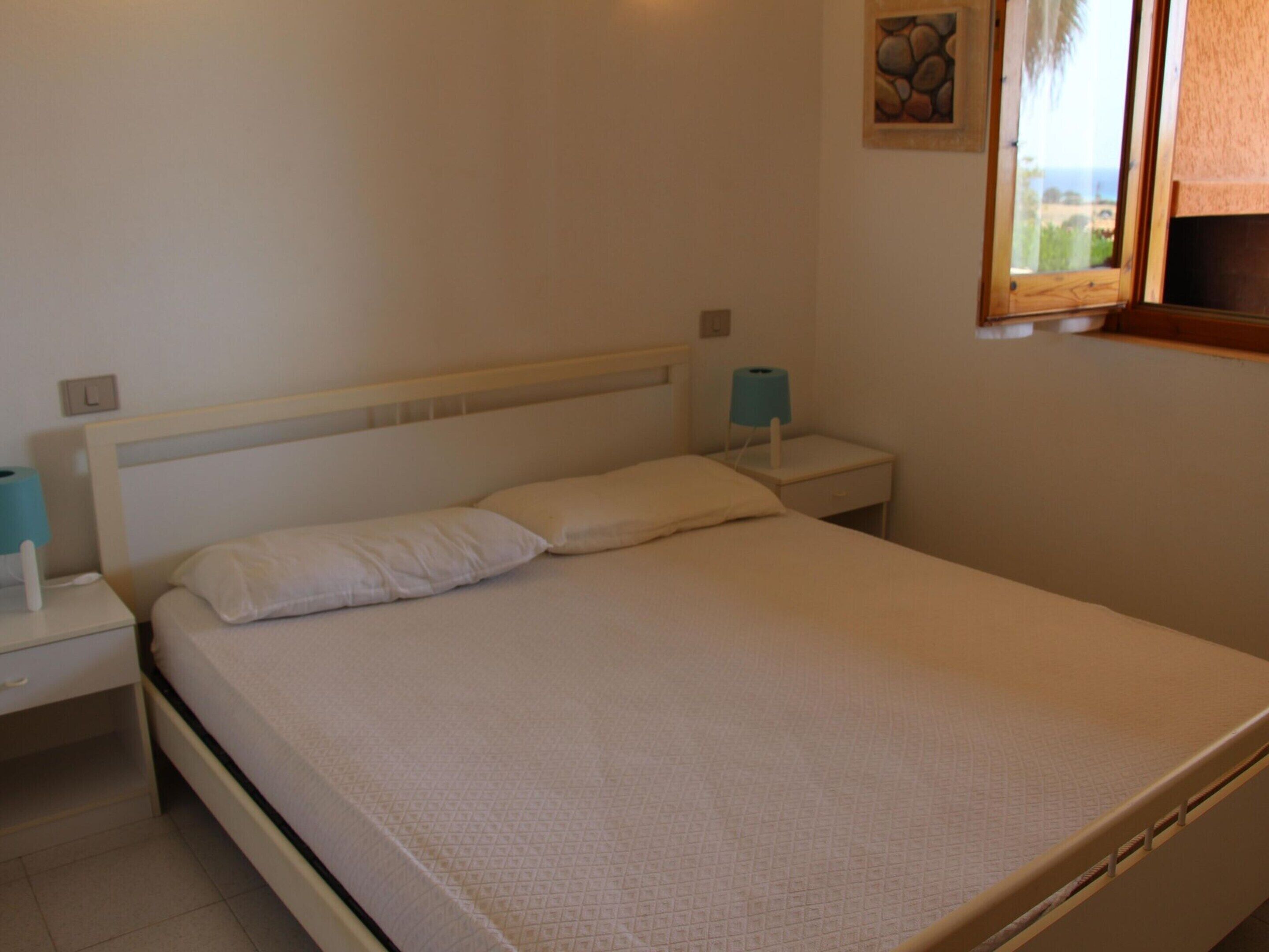 Habitación individual con una cama de soltera, cabecera blanca, mesita de noche con lámpara y ventana con vistas al exterior.