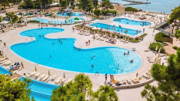 A large outdoor swimming pool complex with multiple pools and sunbathing areas, surrounded by trees and a view of the sea. - Family Fun at Zaton Resort (Nin)