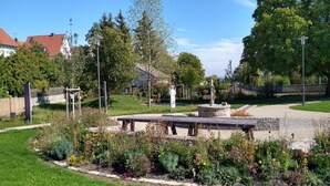En park med en central stenbänk omgiven av blomsterrabatter och träd.