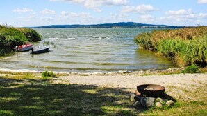 En vy över en sjö med en båt som ligger förtöjd nära stranden, en eldstad på gräsmattan och en avlägsen utsikt över kullar under en delvis molnig himmel.