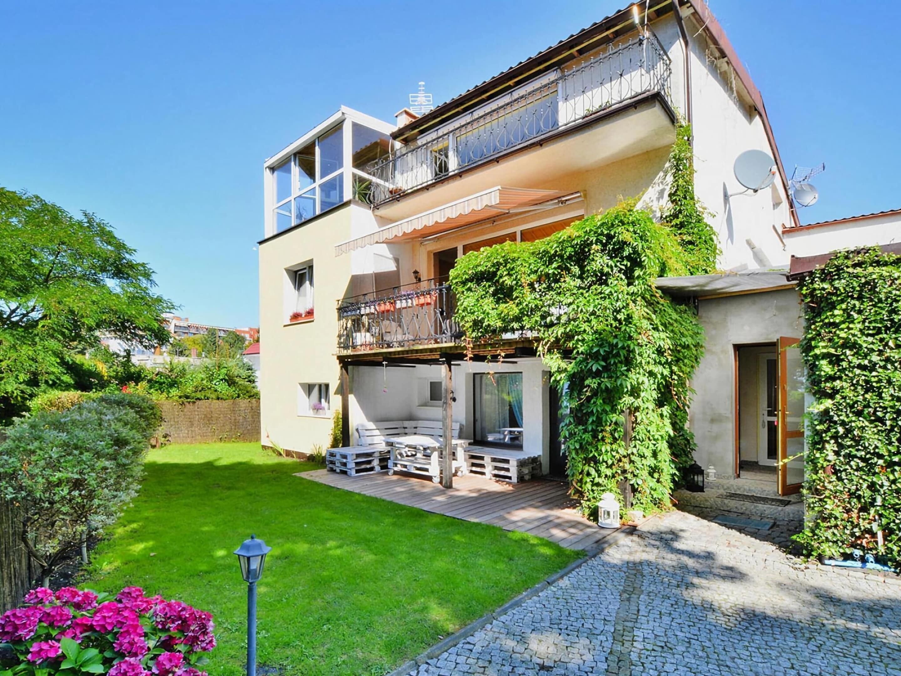 A two-story house with a balcony, a garden, and a paved driveway.
