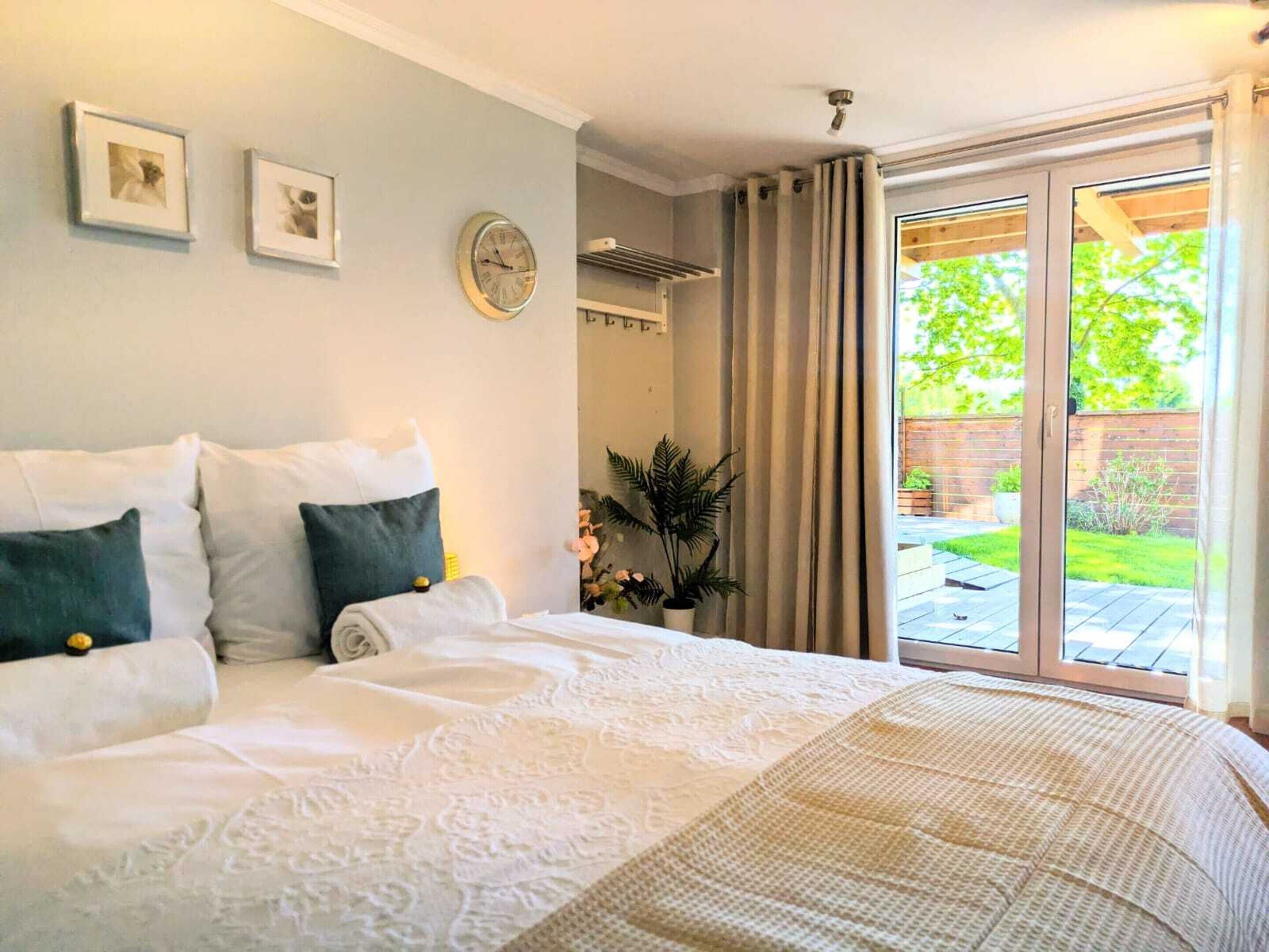 A bedroom with a large bed, a wall clock, framed pictures, and a sliding glass door to a garden.