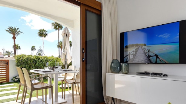 Ein modernes Wohnzimmer mit Fernseher, Esstisch und Stühlen. Ein Schiebeglasfenster führt zu einem Balkon mit Blick auf das Meer und Palmen.
