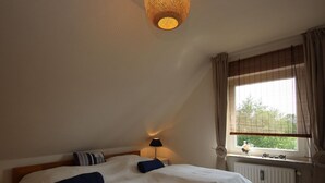 A bedroom with a large bed, a bedside table with a lamp, a window with curtains, and a radiator. - House in Dornumersiel Near Sandy Beach (Dornum)