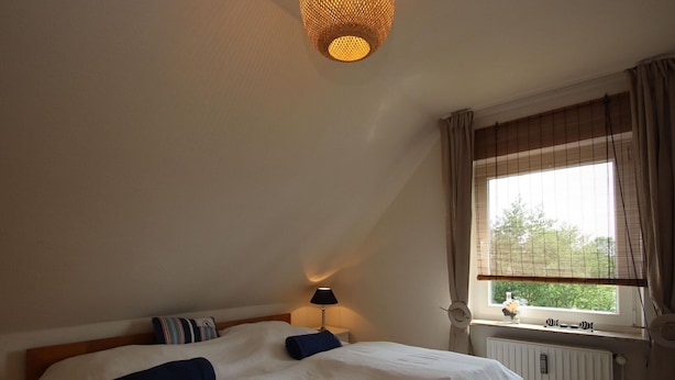 A bedroom with a large bed, a bedside table with a lamp, a window with curtains, and a radiator. - Doppelhaushalfte in Dornumersiel (Dornum)
