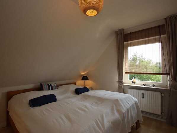 A bedroom with a large bed, a bedside table with a lamp, a window with curtains, and a radiator. - Doppelhaushalfte in Dornumersiel (Dornum)