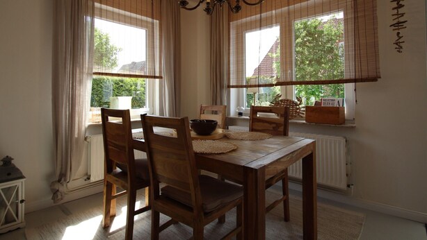 A dining room with a wooden table, chairs, a chandelier, and large windows with blinds. - Doppelhaushalfte in Dornumersiel (Dornum)